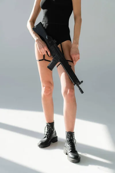 cropped view of woman in leather boots holding rifle on gray background with shadows