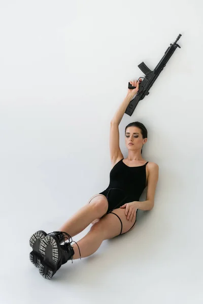 穿着黑色紧身衣和靴子的性感女人 手高高地拿着来福枪 坐在灰色的椅子上 — 图库照片