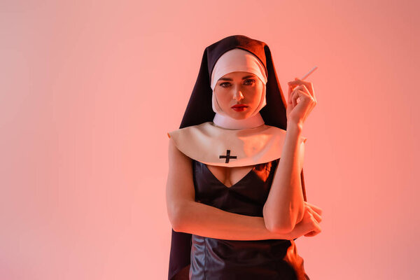 sexy nun in leather dress looking at camera while holding cigarette isolated on pink