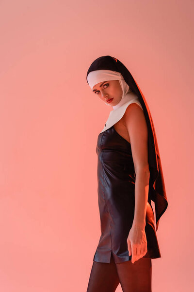 young nun in sexy dress looking at camera while standing isolated on pink