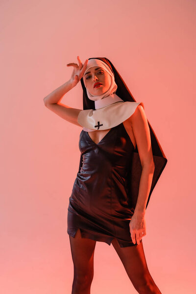 sensual nun looking at camera while holding hand near head isolated on pink