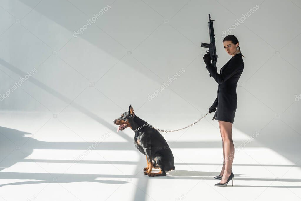 confident woman with rifle near doberman on chain leash on grey background with shadows