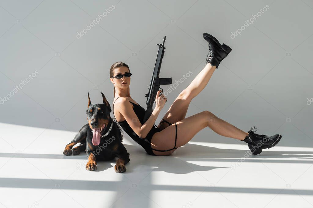 young woman in black bodysuit and sunglasses posing with rifle near doberman on grey background with shadows