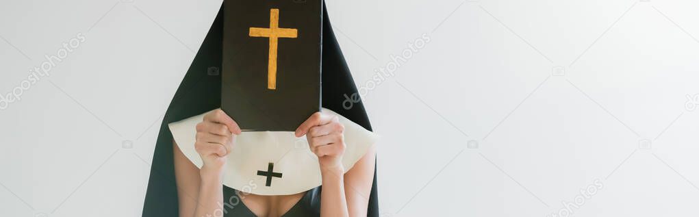 cropped view of sexy nun obscuring face with bible isolated on grey, banner