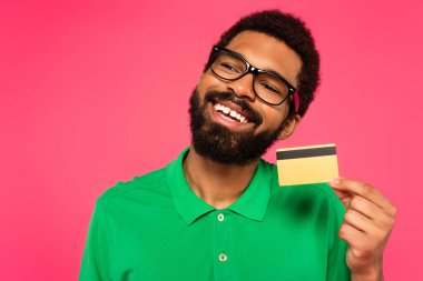 pleased african american man in glasses holding credit card isolated on pink clipart