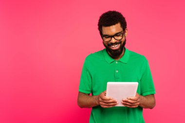 Gözlüklü ve yeşil polo tişörtlü mutlu Afro-Amerikalı adam pembe tablet kullanıyor. 