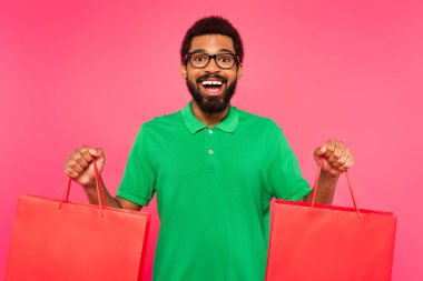 Gözlüklü ve yeşil polo tişörtlü mutlu Afro-Amerikan adam pembe üzerinde izole edilmiş alışveriş torbaları tutuyor.