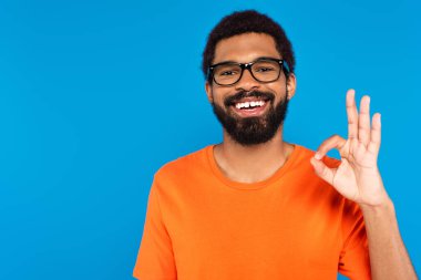 happy african american man in glasses showing ok sign isolated on blue clipart