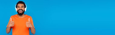 Kablosuz kulaklıklı neşeli Afrikalı Amerikalı adam mavi pankartta izole bir şekilde baş parmaklarını gösterirken müzik dinliyor.