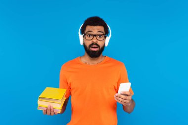 Kafası karışmış, kablosuz kulaklıklı, elinde kitap ve akıllı telefon tutan Afro-Amerikan bir adam.