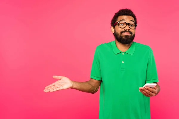 puzzled african american man in green t-shirt holding smartphone isolated on pink