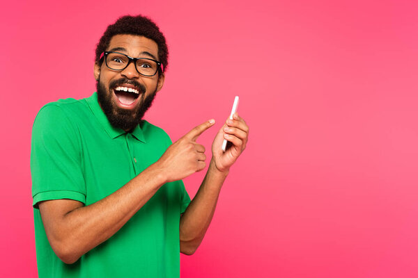 amazed african american man in green t-shirt pointing at smartphone isolated on pink
