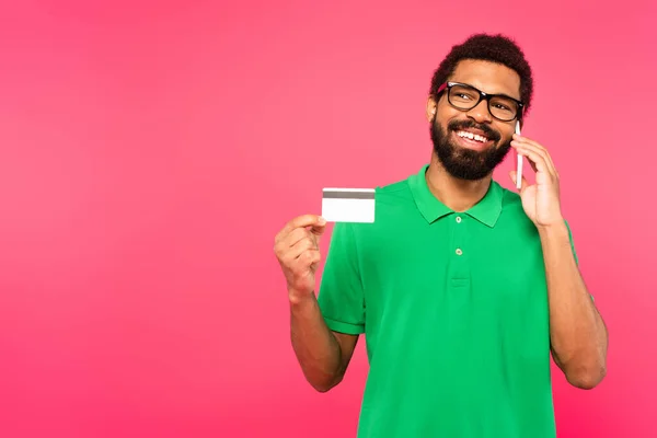 戴眼镜的快乐的非洲裔美国人 一边用智能手机说话 一边拿着用粉红隔开的信用卡 — 图库照片