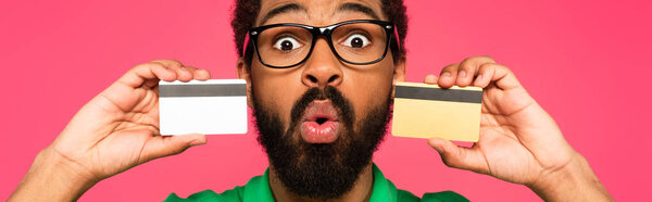 shocked african american man in glasses holding credit cards isolated on pink, banner
