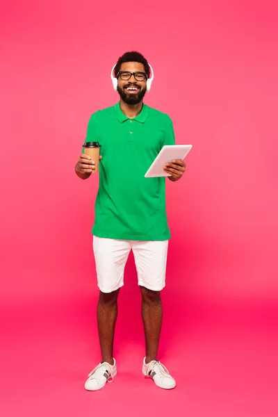 ピンクの紙コップとデジタルタブレットを持っている眼鏡やヘッドフォンの明るいアフリカ系アメリカ人男性の完全な長さ — ストック写真