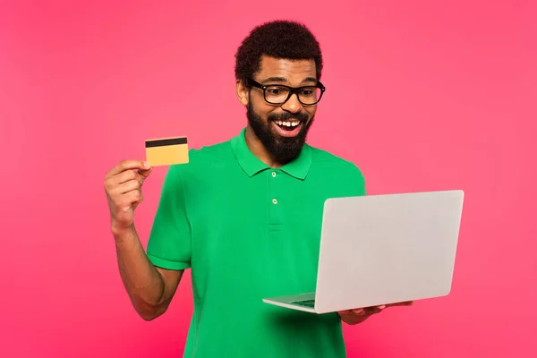 幸せなアフリカ系アメリカ人男性の眼鏡と緑のポロシャツノートパソコンとクレジットカードをピンクに隔離 — ストック写真