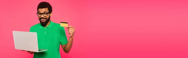 幸せなアフリカ系アメリカ人男性の眼鏡と緑のポロシャツノートパソコンとクレジットカードを持ってピンク バナーに隔離された — ストック写真