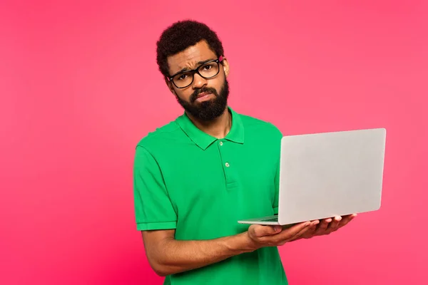 身穿眼镜和绿色马球衫的非洲裔美国人很不高兴 他拿着用粉色隔离的笔记本电脑 — 图库照片