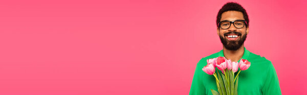 веселый африканский американец в очках и зеленой рубашке в поло с тюльпанами, изолированными на розовом, баннер