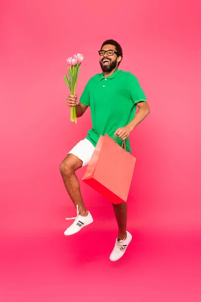 幸せなアフリカ系アメリカ人男性の完全な長さで眼鏡をかけてショッピングバッグとチューリップの花束ピンク — ストック写真