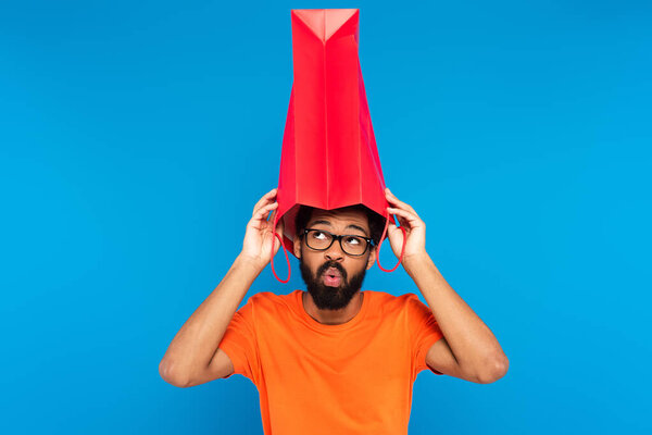 surprised african american man in glasses holding shopping bag above head isolated on blue 