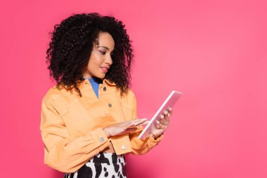 Kıvırcık Afrikalı Amerikalı kadın pembe üzerine izole edilmiş dijital tablet kullanıyor. 