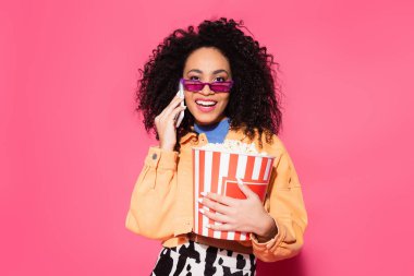 Güneş gözlüklü mutlu Afro-Amerikalı kadın elinde patlamış mısır kovası ve pembe telefonda akıllı telefonla konuşuyor. 