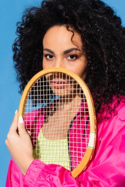Kıvırcık Afrikalı Amerikalı kadın tenis raketiyle poz verirken mavi kameraya bakıyor.