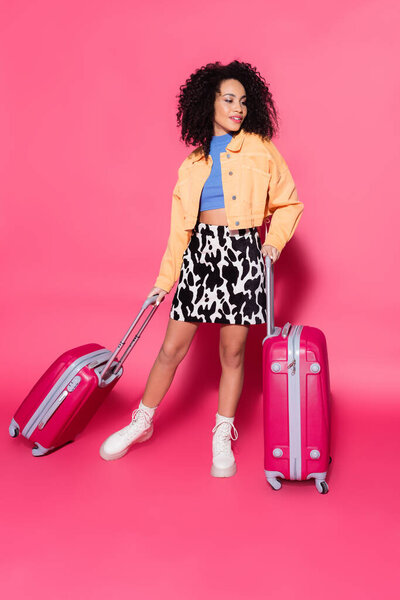 full length of stylish african american woman posing with suitcases on pink 