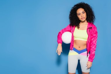curly african american woman in pink jacket and crop top standing with volleyball on blue clipart