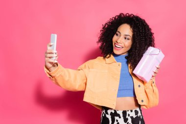 Mutlu Afro-Amerikan kadın ceketini elinde tutarken pembedeki akıllı telefondan selfie çekiyor. 