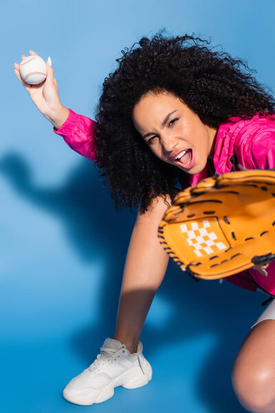 excited african american woman in leather glove holding baseball on blue
