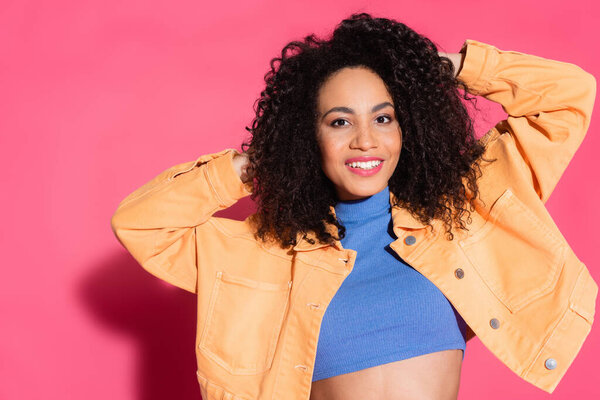 smiling african american woman in jacket and blue crop top posing on pink 