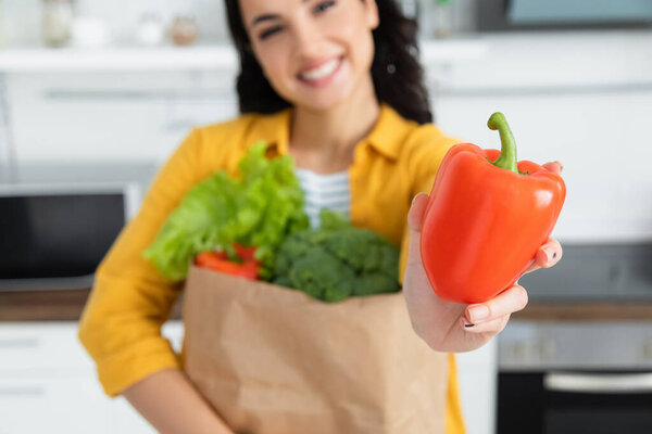 blurred and happy brunette woman holding paper bag with groceries and bell pepper 