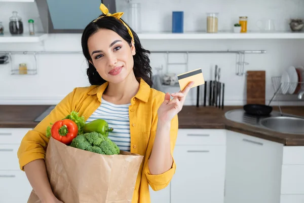 smiling brunette woman holding paper bag with groceries and credit card