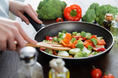partial view of woman mixing sliced vegetables on frying pan with spatula  clipart
