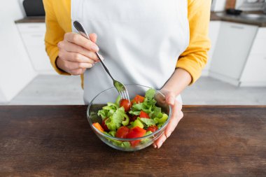 Önlüklü genç bir kadının elinde çatalla kasede salata tutarken çekilmiş görüntüsü.