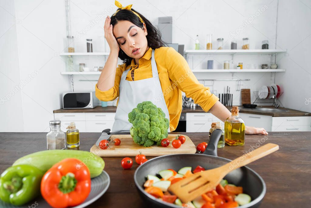tired young woman in apron looking at vegetables near frying pan in kitchen 