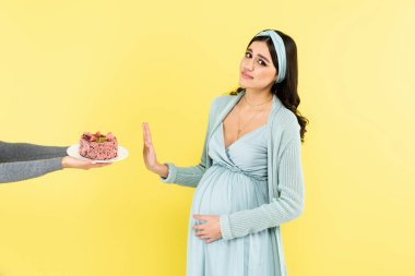 Hamile kadın kameraya bakarken dur işareti gösteriyor. Sarı renkte izole edilmiş pasta yanında.