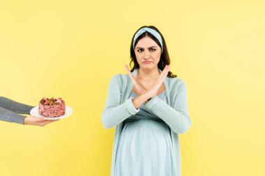 disgusted, pregnant woman showing reject gesture near piece of cake isolated on yellow clipart