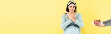 displeased pregnant woman showing reject gesture near cake isolated on yellow, banner clipart