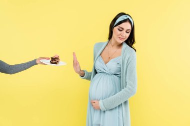 young pregnant woman showing reject gesture near sweet dessert isolated on yellow clipart