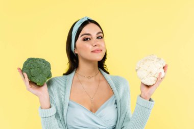 young woman smiling at camera while holding cauliflower and broccoli isolated on yellow clipart