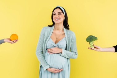 Genç hamile bir kadın taze portakal ve brokoli yanında gülümsüyor.