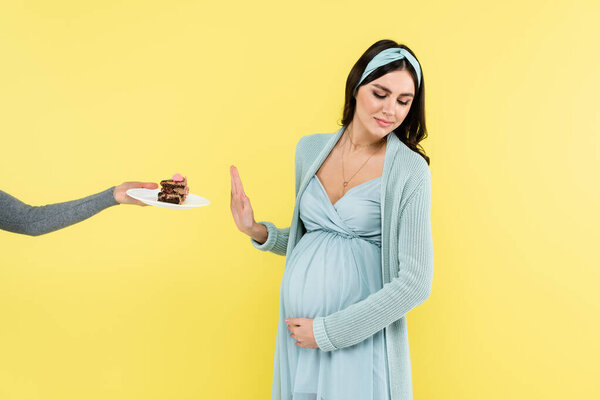 young pregnant woman showing reject gesture near sweet dessert isolated on yellow