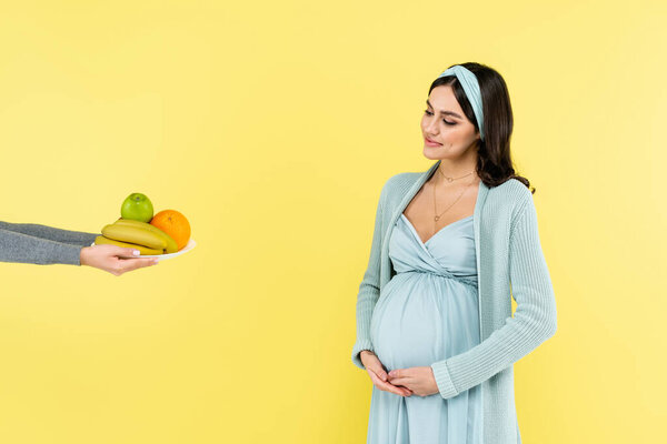 smiling pregnant woman touching belly while looking at fresh fruits isolated on yellow