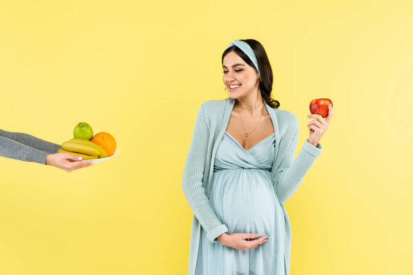 cheerful pregnant woman holding ripe apple while looking at fresh fruits isolated on yellow