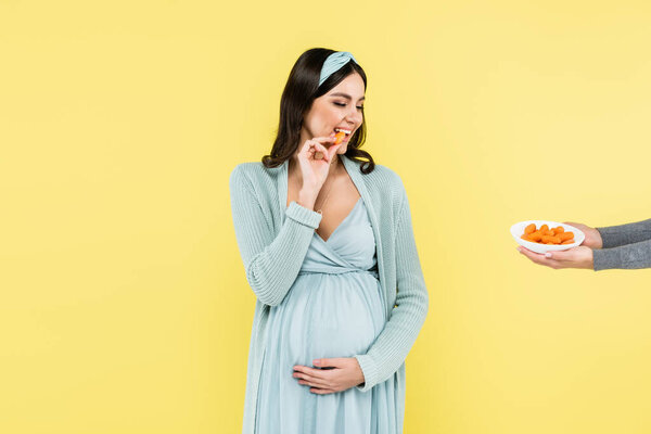 cheerful pregnant woman looking at bowl with fresh carrot isolated on yellow