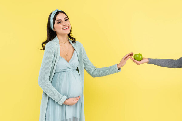 happy pregnant woman smiling at camera while taking fresh apple isolated on yellow