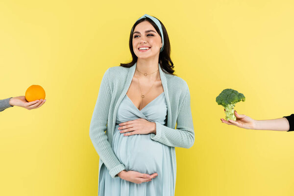 young pregnant woman smiling near fresh orange and broccoli isolated on yellow